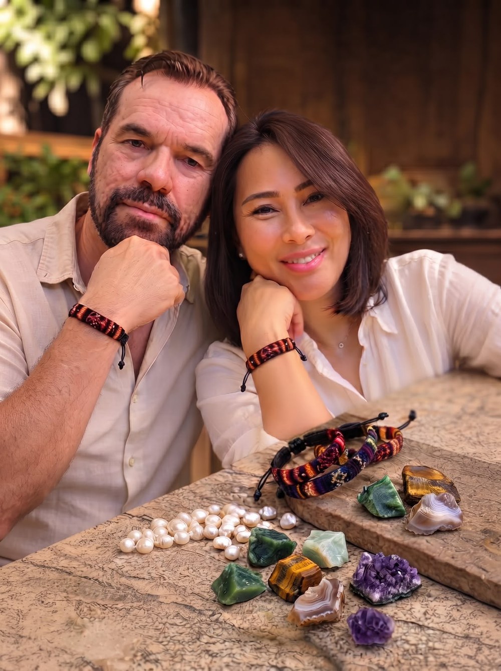A man and a woman sitting close together at a stone table, both wearing woven bracelets, with pearls, woven bracelets and raw gemstones laid out in front of them