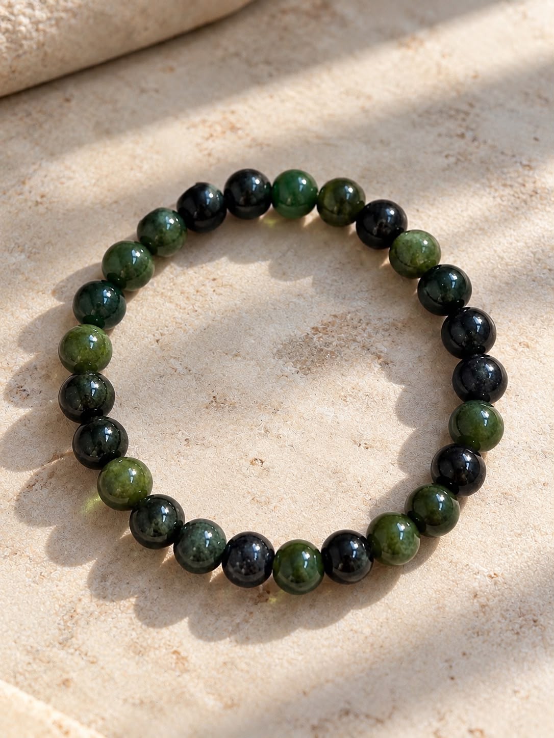 A bracelet of round green and black jade beads on pale stone in sunlight